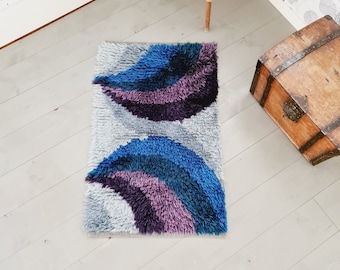 May include: A gray, blue, and purple shag rug with a wavy, abstract design. The rug is on a white wood floor.