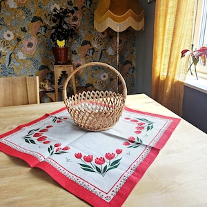 May include: A woven wicker basket with a handle sits on a white tablecloth with a red border and red tulip floral design. The basket is on a wooden table. The background features floral wallpaper and a yellow lamp.
