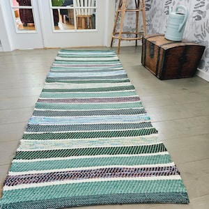 Puede incluir: Una alfombra tejida a rayas verdes, azules y blancas con una sutil raya morada. La alfombra está hecha de fibra natural y tiene un aspecto rústico y hecho a mano.