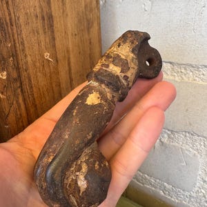 May include: A heavy, weathered metal door knocker with a dark, rusted finish and patches of light-colored oxidation. The object features a curved, elongated handle design with a rounded base and a circular mounting hole at the top section.