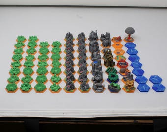 May include: A collection of miniature board game pieces arranged in rows on hexagonal bases. The set includes green foliage tiles, grey rocky terrain, grey industrial structures, and blue water tiles, along with several multi-colored special tokens.