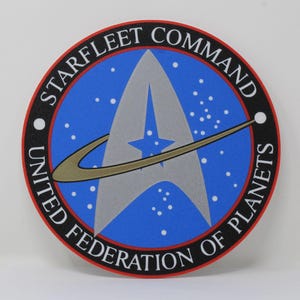 Könnte beinhalten: Rundes Emblem mit der Aufschrift "STARFLEET COMMAND" und "UNITED FEDERATION OF PLANETS". Es hat einen blauen Hintergrund mit weißen Sternen, eine silberne "A"-Form mit einem blauen Stern und einen goldenen Ring.