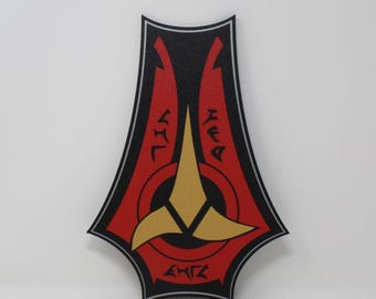 May include: A shield-shaped wall plaque featuring a black background with a red central area and a gold three-pointed emblem. The design includes silver border lines and black alien-style lettering above and below the central gold symbol.