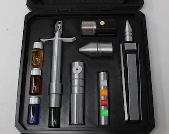 May include: A black carrying case containing a collection of metallic and plastic tools. The set includes a silver syringe-like device, three small vials with orange, red, and blue liquid, a silver cone, a black cylinder with a yellow button, and a rectangular silver and black tool with four colored square buttons.