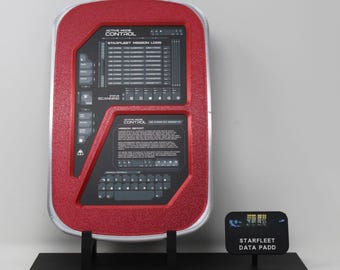 May include: A red and silver rectangular Starfleet data PADD prop featuring a black screen with white text including Active Mode Control, Starfleet Mission Logs, and a keyboard layout. It is mounted on a black stand with a small matching sign.