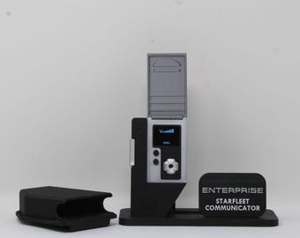 May include: A silver and black Starfleet Communicator replica with an open lid, a digital display showing signal bars, and a central dial. It sits on a black stand next to a black case. The base features a plaque reading ENTERPRISE STARFLEET COMMUNICATOR.