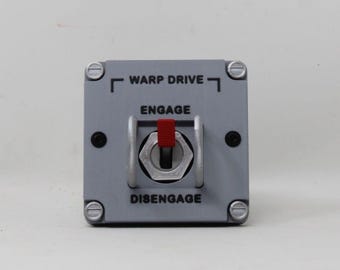 May include: A square gray control panel featuring a central toggle switch with a red tip. The panel is labeled with the text WARP DRIVE at the top, and ENGAGE and DISENGAGE above and below the switch. It is secured by four silver-colored screws.