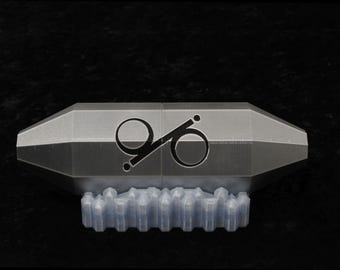 May include: A metallic, silver-toned, hexagonal-shaped object with tapered ends, featuring a black, stylized circular logo printed on the center. The object is supported by a translucent, light-colored, multi-segmented base stand.