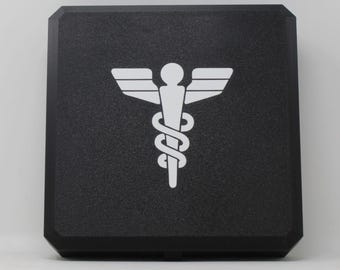 May include: A square black box with a textured surface featuring a white caduceus symbol printed in the center. The medical emblem consists of a central staff with two wings at the top and a snake coiled around the lower portion of the staff.