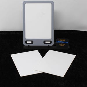May include: A gray and white Star Trek Lower Decks PADD prop with two white square cards. The PADD has a rectangular screen and two black buttons. The Starfleet PADD logo is visible on a black base.