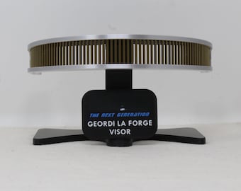 May include: A curved, metallic visor with a bronze-colored slatted front and silver trim, mounted on a black stand. A black plaque on the stand features the text THE NEXT GENERATION GEORDI LA FORGE VISOR in white and blue lettering.