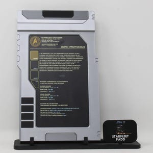 May include: A silver Starfleet PADD prop featuring a black screen with white text titled Gorn Protocols, Starfleet Database, and Phaser Harmonics Adjustments. The device is mounted on a black stand with a small sign labeled Starfleet PADD.