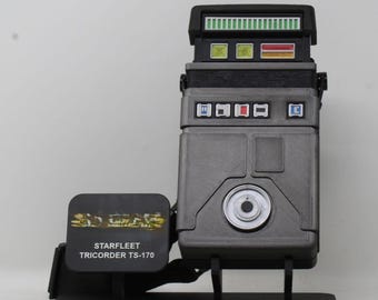 May include: A grey Starfleet Tricorder TS-170 replica mounted on a black stand. The device features a top section with green, yellow, and red indicator lights, a central body with four small square buttons, and a circular silver lens at the bottom.