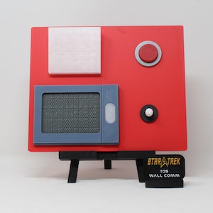 May include: A red Star Trek TOS Wall Comm prop replica. The prop features a square red face with a silver square, a blue rectangular speaker, and two circular buttons. The prop is mounted on a black stand with the Star Trek logo.