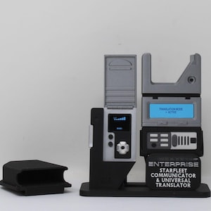 May include: A Star Trek Enterprise Starfleet Communicator and Universal Translator device with a gray and black finish. The unit features a digital display showing "TRANSLATION MODE ACTIVE" and a separate small screen with signal bars.
