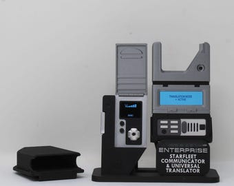 May include: A Star Trek Enterprise Starfleet Communicator and Universal Translator device with a gray and black finish. The unit features a digital display showing "TRANSLATION MODE ACTIVE" and a separate small screen with signal bars.