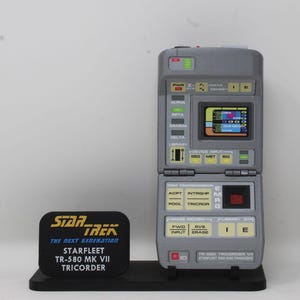May include: A grey Star Trek TR-580 MK VII Tricorder prop replica. The device features a flip-up screen and various buttons labelled "Alpha," "Beta," and "Gamma." A black base displays the Star Trek logo and device details.