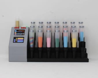 May include: A gray console with a digital display showing the text "COMPOUND DYE TANK" and numerical data, positioned next to a black rack holding seven clear cylindrical tubes. Each tube contains a different colored powder or liquid substance.