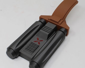 May include: A matte black dual-barrel prop with a textured brown handle extension. The central section features a series of horizontal ridges and a red cross-shaped emblem. The object has a geometric, industrial design with cylindrical barrels.