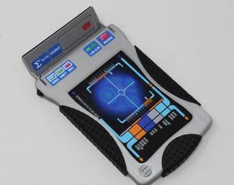 May include: A silver handheld electronic device with black textured side grips and a screen displaying a blue crosshair graphic. The device features buttons labeled SYS COM, LIB A/B, EMRG, and GEO MET BIO, with text reading BIOMOLECULAR SCAN 483R.