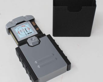 May include: A rectangular electronic device with a sliding grey cover and a black base, featuring a screen displaying a circular radar-style graphic with red and black markings. A separate black rectangular storage case is positioned nearby.