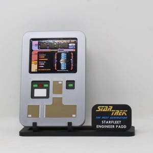 May include: A silver Star Trek: The Next Generation Starfleet Engineer PADD prop replica. The PADD features a screen with colorful graphics and buttons. The base has the Star Trek logo and text.
