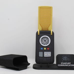 May include: A black and gold Starfleet communicator device with a flip-up antenna, a circular speaker grille, three coloured buttons, and two silver control knobs. It sits on a black stand next to a black carrying case and a sign reading Starfleet Communicator.