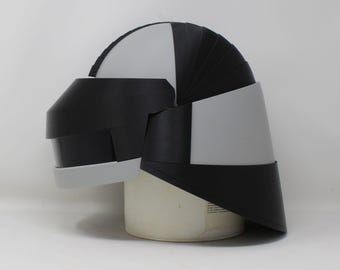 May include: A futuristic helmet featuring a segmented design with alternating matte black and light gray panels. The helmet has a smooth, curved dome shape with a prominent visor section and a flared back, constructed from rigid, layered components.