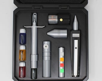 May include: A black storage case containing a collection of silver, black, and metallic tools. The set includes a long silver syringe-like tool, three small glass vials in blue, amber, and red, a silver cylinder, a silver cone, and a black device.
