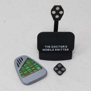 以下が含まれることがあります： 「THE DOCTOR'S MOBILE EMITTER」という文字が入った黒いディスプレイスタンド。垂直なアームの先端には4つの銀色の磁石が付いた円形のパーツがある。その横には、緑色のグリッド模様が入った灰色の盾型のデバイスと、4つの銀色の磁石が付いた小さな黒いひし形のパーツが置かれている。