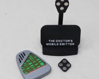 May include: A black display stand with the text THE DOCTOR'S MOBILE EMITTER, featuring a vertical arm with a circular top containing four silver magnets. Beside it lies a gray, shield-shaped device with a green grid pattern and a small black diamond-shaped piece with four silver magnets.