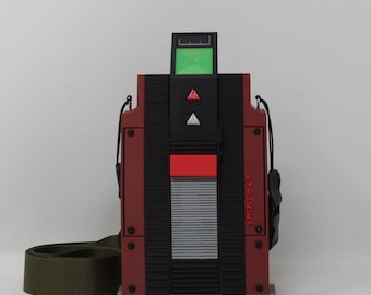 May include: A rectangular maroon bag with black vertical textured panels, a green screen display at the top, and red and white triangular accents. It features a silver horizontal slatted section, black side hardware, and an olive green shoulder strap.