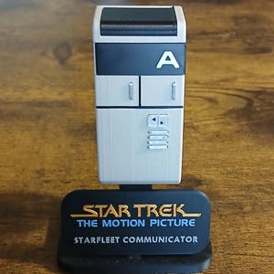 Puede incluir: Una réplica del comunicador Starfleet de Star Trek: La película. El dispositivo es principalmente blanco con una parte superior negra y presenta la letra "A". La base es negra con texto dorado que dice "STAR TREK THE MOTION PICTURE STARFLEET COMMUNICATOR."