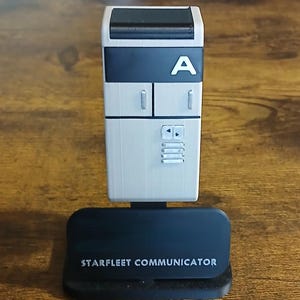 Puede incluir: Una réplica rectangular de comunicador Starfleet en gris claro y negro, con una sección superior negra, una letra A blanca y botones de control plateados. El dispositivo está montado sobre una base negra con el texto STARFLEET COMMUNICATOR.