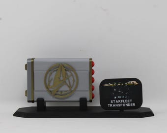May include: A silver rectangular Starfleet transponder replica featuring a gold-colored emblem on the front and five red circular accents on the side. The device is mounted on a black display stand with a small black plaque labeled STARFLEET TRANSPONDER.