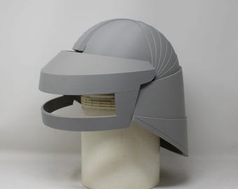 May include: A matte gray helmet featuring a rounded dome, a wide visor section, and a curved neck guard. The helmet has a layered, segmented design on the sides and is displayed on a cylindrical stand, showcasing a smooth, uniform finish.
