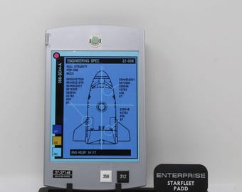 May include: A silver rectangular electronic device with a blue screen displaying a schematic of a spacecraft, technical data, and the text ENGINEERING SPEC 22-008. It features buttons labeled DAT, LOC, REC, 356, and 312, resting on a black stand with a sign reading ENTERPRISE STARFLEET PADD.