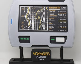May include: A silver rectangular Star Trek Voyager PADD prop featuring a screen displaying human anatomy diagrams and text including Human Anatomy-03.7329. The device is mounted on a black stand with a plaque reading Voyager Starfleet PADD.