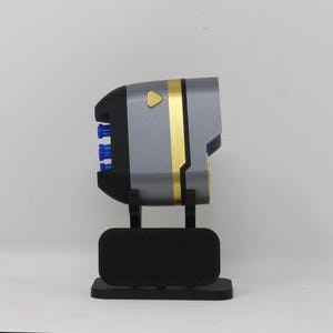Puede incluir: Un dispositivo de utilería gris y negro con una banda dorada y un pequeño botón triangular dorado en el lateral. El objeto presenta tres componentes cilíndricos azules en la parte trasera y está montado sobre un soporte rectangular negro.