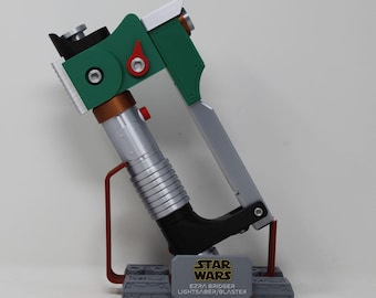 May include: A green, silver, and black Ezra Bridger Lightsaber/Blaster prop from Star Wars. The prop features a green handle with red and silver accents, a silver barrel, and a black grip. The base has the Star Wars logo and text.