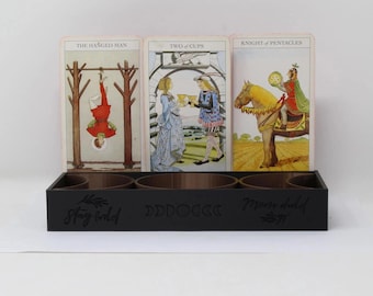 May include: A black wooden tarot card holder with three tarot cards displayed. The cards are "The Hanged Man", "Two of Cups", and "Knight of Pentacles". The holder has the words "Stay Wild" and "Moon Child" on it.