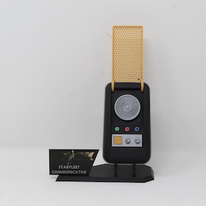 May include: A black Starfleet communicator replica with a yellow perforated flip cover, a circular silver speaker grille, three small colored buttons, and a rectangular control panel. It is mounted on a black display stand with a label reading STARFLEET COMMUNICATOR.