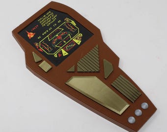 May include: A brown, handheld, sci-fi style prop device featuring a black screen with yellow alien-like text and a central graphic. The device includes various metallic-colored geometric buttons, a ridged panel, and three small circular silver buttons.