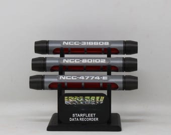 May include: A Starfleet Data Recorder display stand featuring three cylindrical gray and black tubes with red centers. Each tube is labeled with white text: NCC-316608, NCC-80102, and NCC-4774-E. The black base features the text STARFLEET DATA RECORDER.
