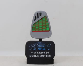 May include: A gray, shield-shaped mobile emitter featuring a green and brown grid pattern with black vents at the top. It is mounted on a black rectangular base with the white text THE DOCTOR'S MOBILE EMITTER printed on the front surface.