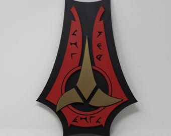 May include: A black, red, and gold wall decoration with a stylized design. The central element is a gold emblem within a red circle, with a three-pointed shape below. Red accents and symbols are on the black background.