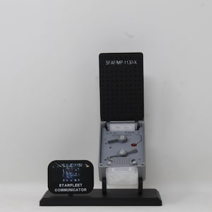 May include: A gray and black Starfleet communicator prop on a black stand. The device features a flip-top lid with the text SFAF/MP-1130-X, a control panel labeled S.F.G TYPE 3, and a small black sign reading STARFLEET COMMUNICATOR in white text.
