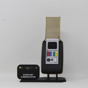 May include: A Starfleet communicator replica featuring a black rectangular base with a gold-colored ribbed antenna section extended upward. The device includes a silver control panel with a black screen, red, blue, green, and yellow buttons, and a black plaque.