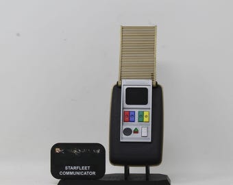 May include: A Starfleet communicator replica featuring a black rectangular base with a gold-colored ribbed antenna section extended upward. The device includes a silver control panel with a black screen, red, blue, green, and yellow buttons, and a black plaque.