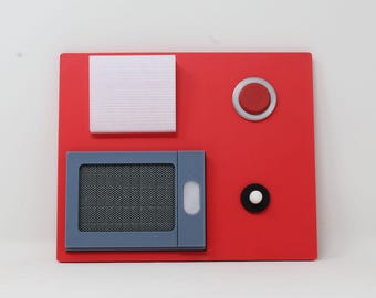 May include: A red rectangular panel with several geometric shapes. It features a square with a textured silver surface, a grey rectangular shape with a mesh screen, a red button, and a black and white button.